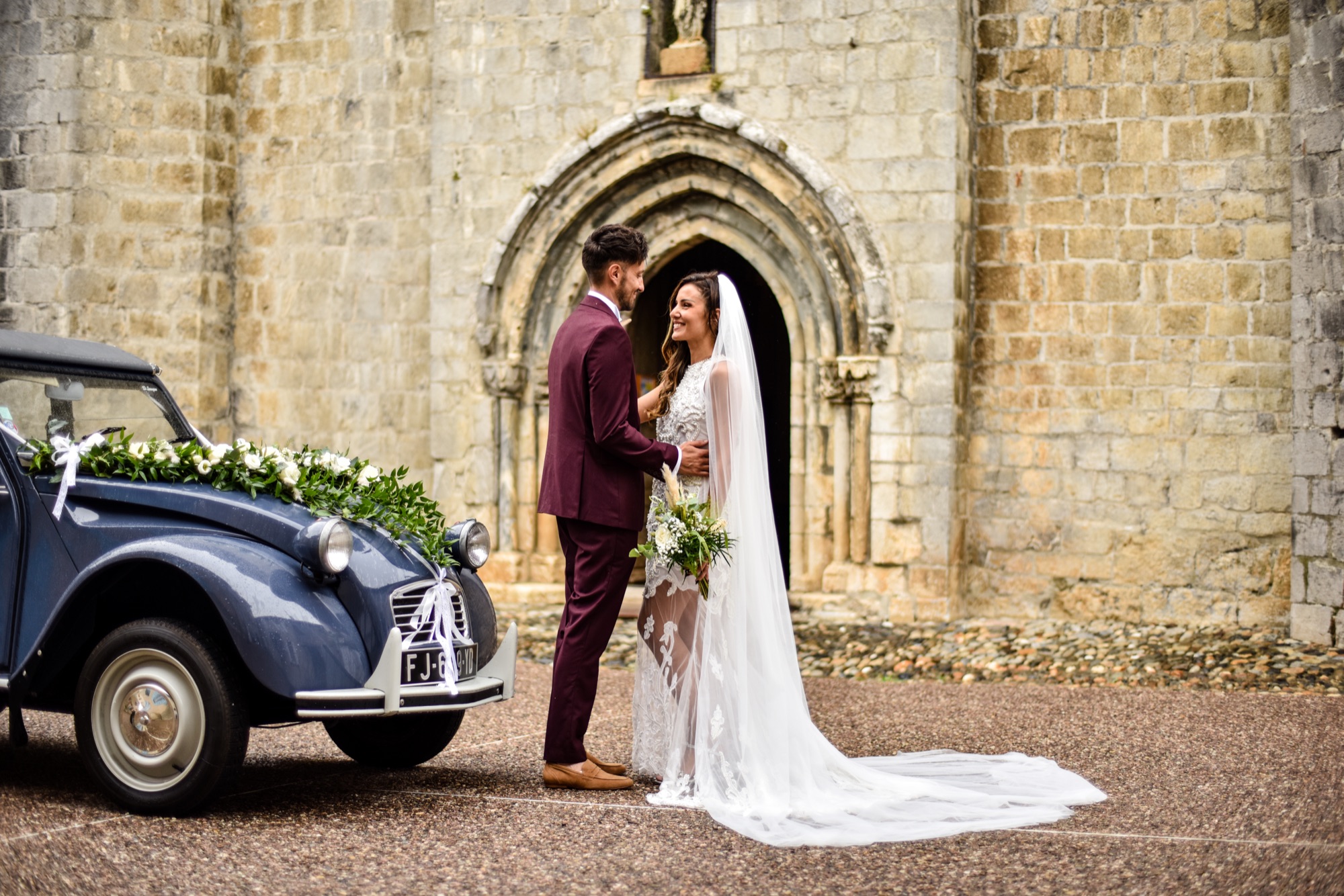 Couple devant une église en pierre avec une 2CV fleurie